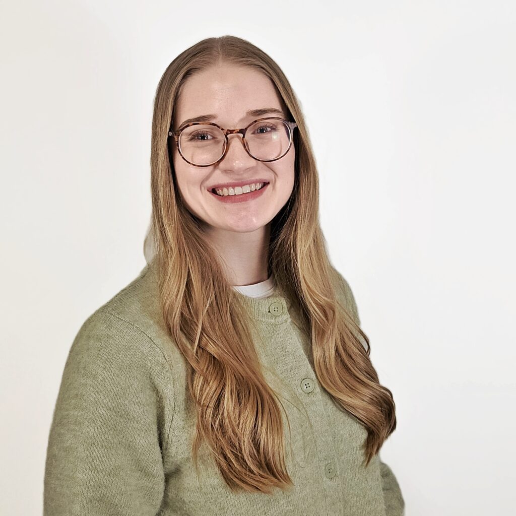 A head shot of Lynn Penner. She has long blonde hair hanging forward over both her shoulders, round-framed brown glasses, and is wearing a light green buttoned cardigan.