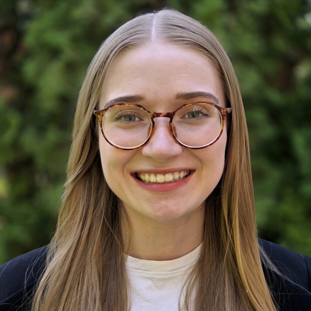 A headshot photo of Lynn Penner. Lynn has long, straight blonde hair, round-framed brown glasses, and is smiling while looking straight at the camera. She is wearing a black blazer over a white T-shirt.